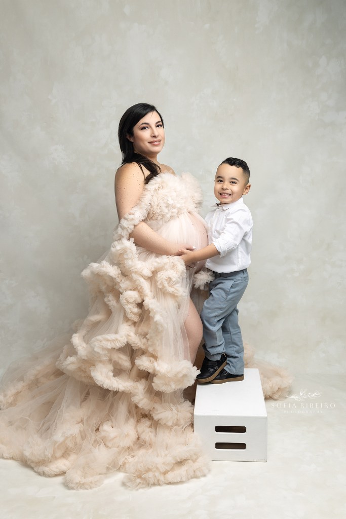 mom holds her little son's hands while dressed in a cream ruffled gown exposing her belly during a maternity session in nj
