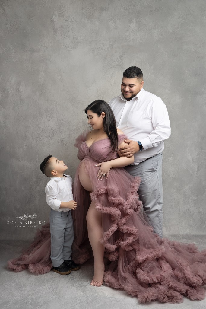 mom holds her little son's hands while her husband stands behind her dressed in a pink maternity gown