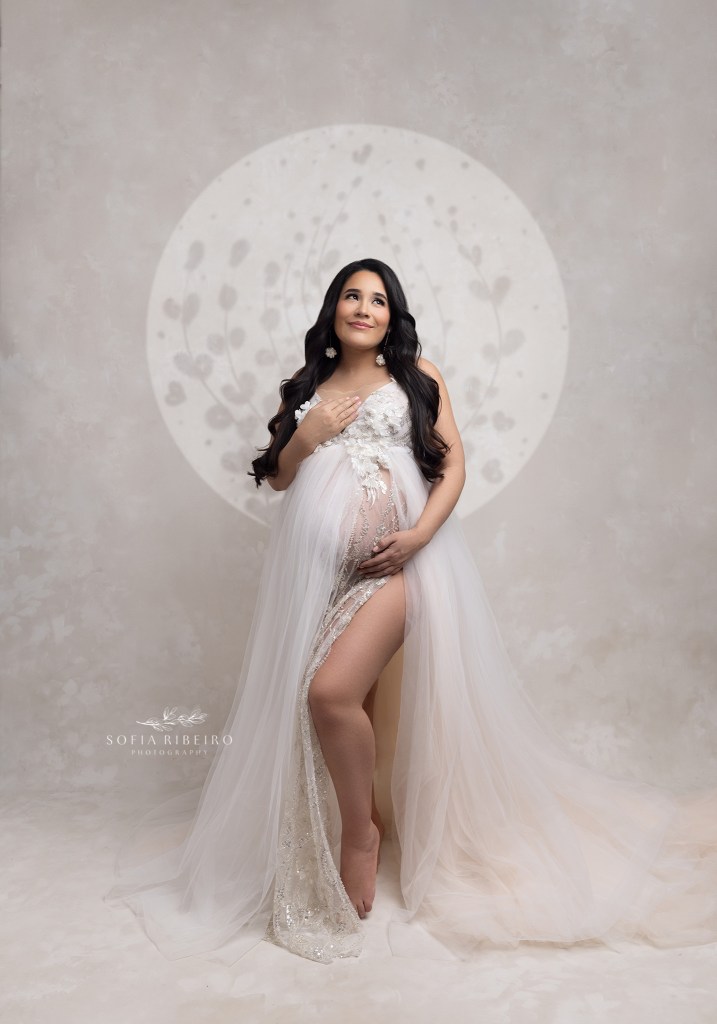 mom poses in a white beaded dress cradling her bump with a halo outline surrounding her