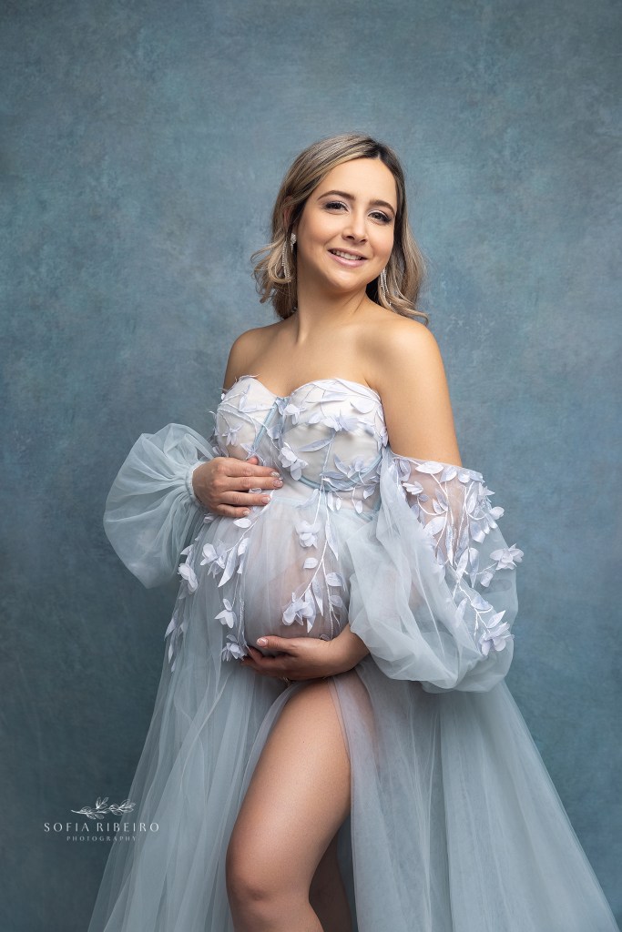 mom smiles at the camera wearing a baby blue floral dress during a maternity photo session