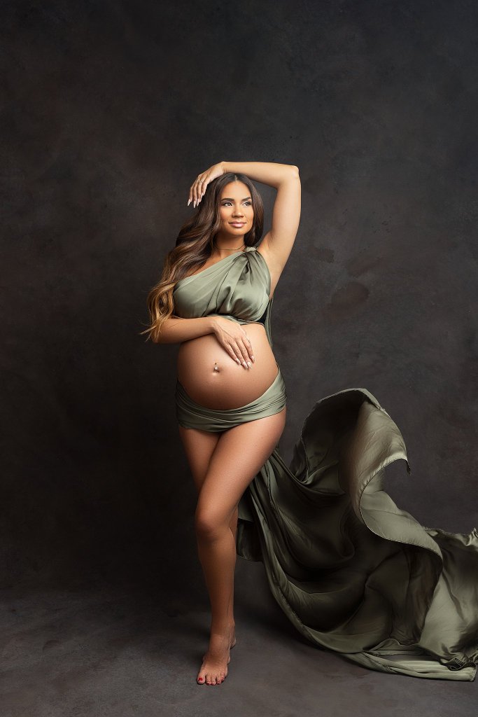 A pregnant woman stands in a studio wearing only a green sheet with it blowing in the wind after a hoboken prenatal massage
