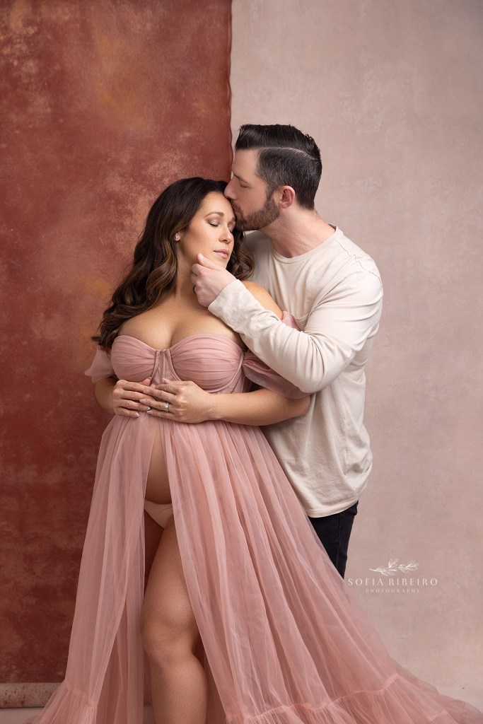 dad cradles moms face while kissing her on the forehead, standing in front of two layered pink backdrops in a nj maternity studio
