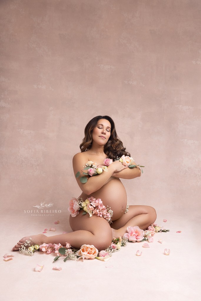 mom poses nude, with arms crossed in front of her, draped in flower highlighting her belly during a nj maternity session