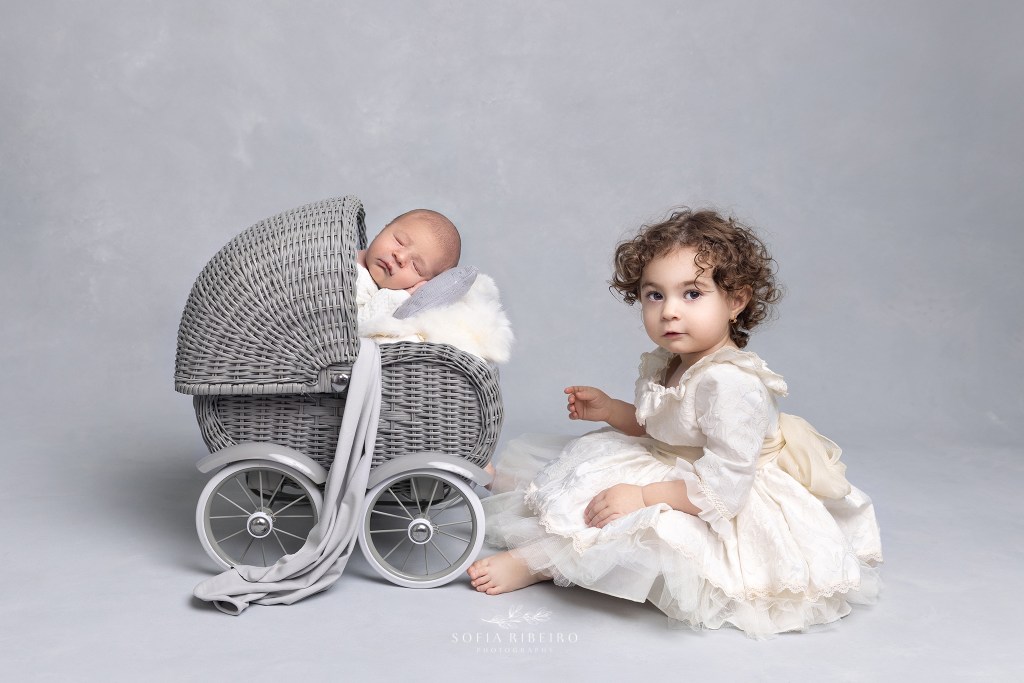 baby is posed in a newborn baby prop carriage during his newborn session with his big sister close by