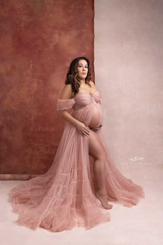 mom looks towards the camera in a pink gown while standing in front of two layered pink backdrops in a nj maternity studio