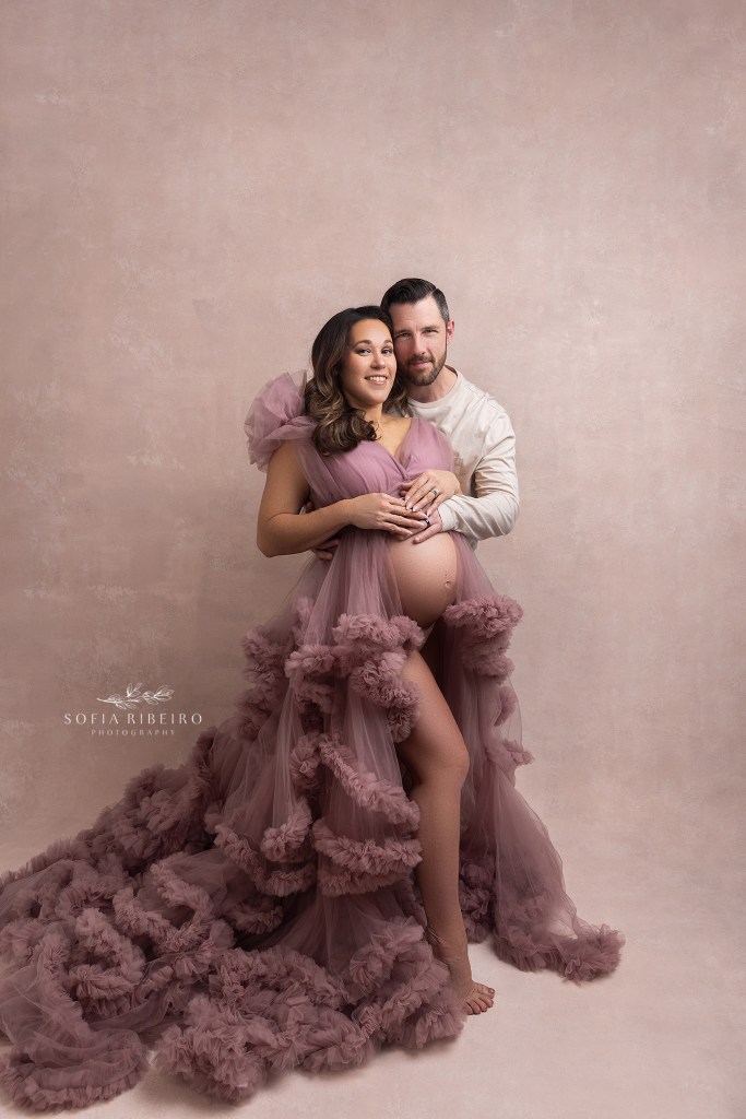 mom poses during a maternity photo session in new jersey against a pink backdrop with dad in a cream shirt