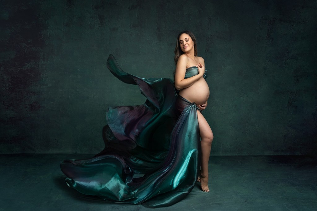 A mother to be stands in a studio wrapped in shiny green fabric that is blowing in the wind behind her after visiting our birthing center