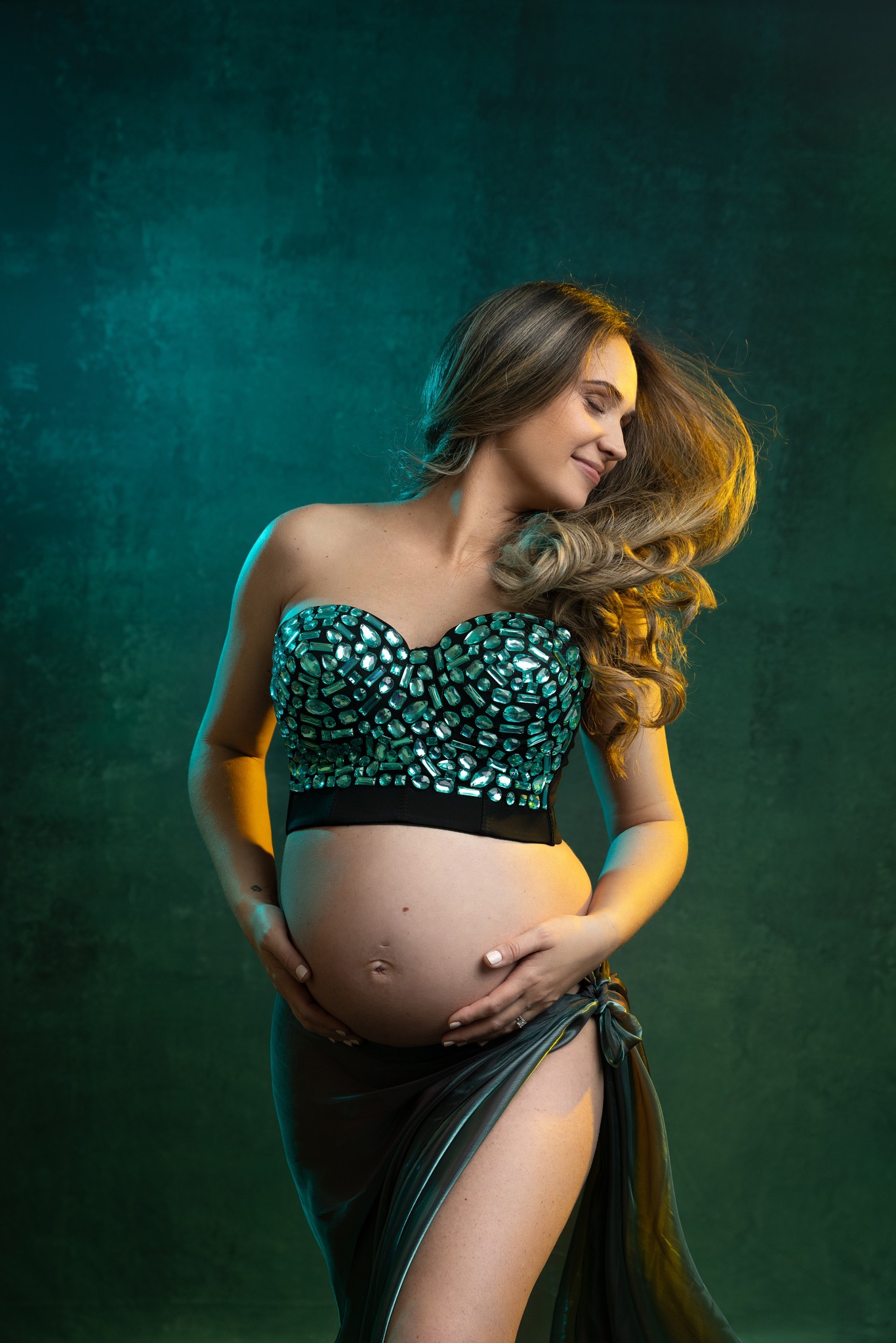 A mom to be stands in a studio holding her bump and flipping her hair behind her after visiting our birthing center