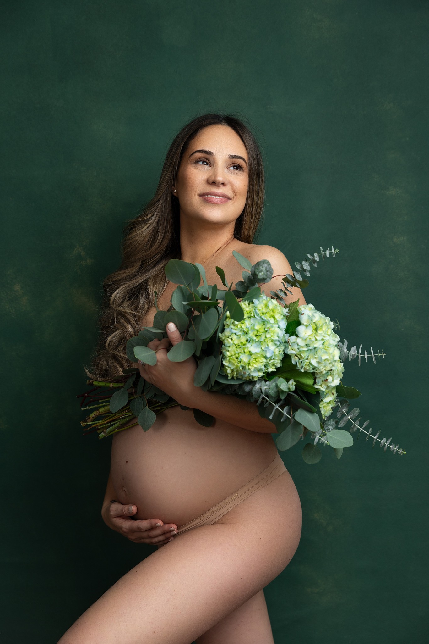 A mom to be stands in a studio smiling while covering with a bouquet of flowers