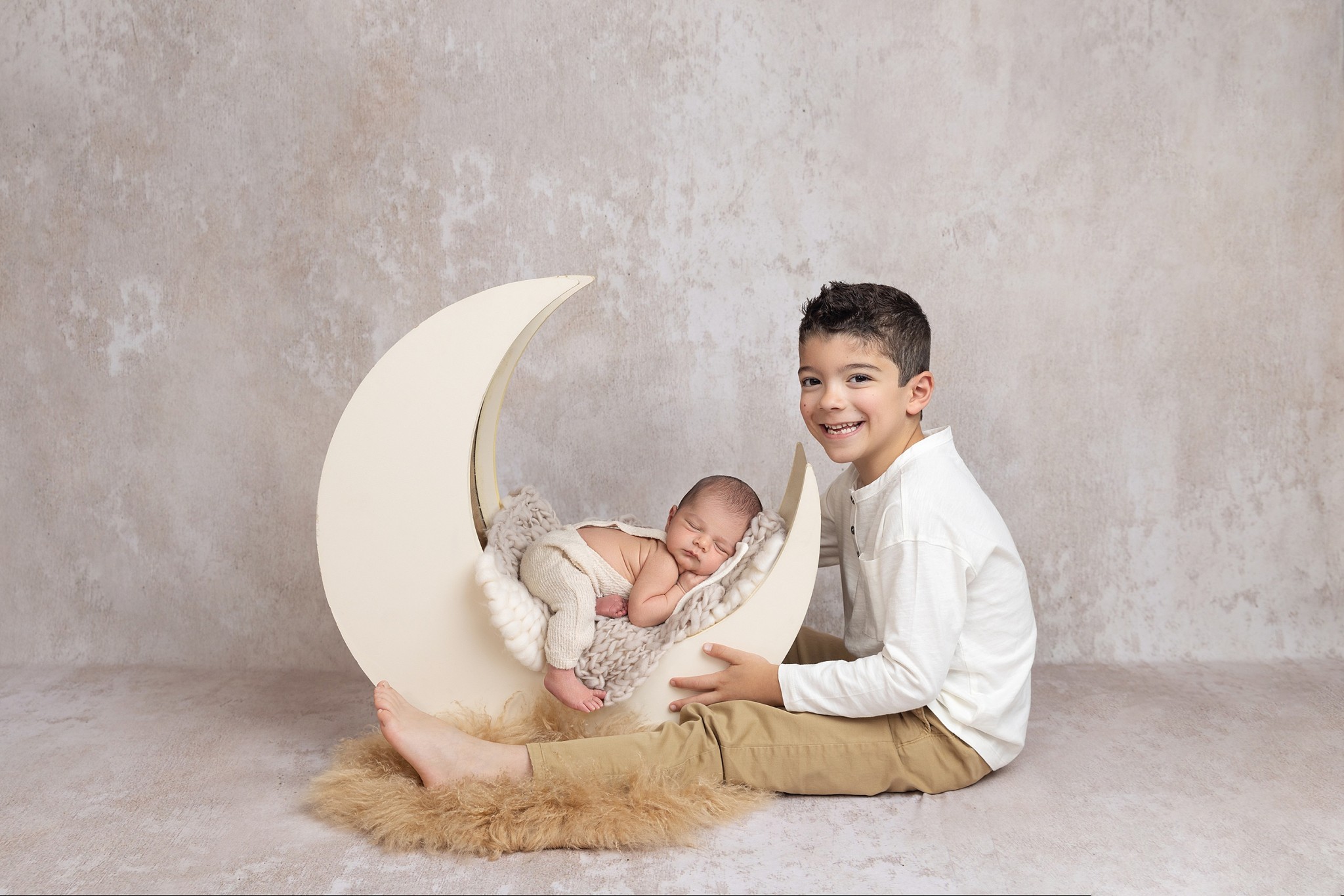 A toddler boy in a white shirt and khakis sits in a studio with his newborn baby sibling sleeping in a moon shape crib after meeting pediatric chiropractors nj