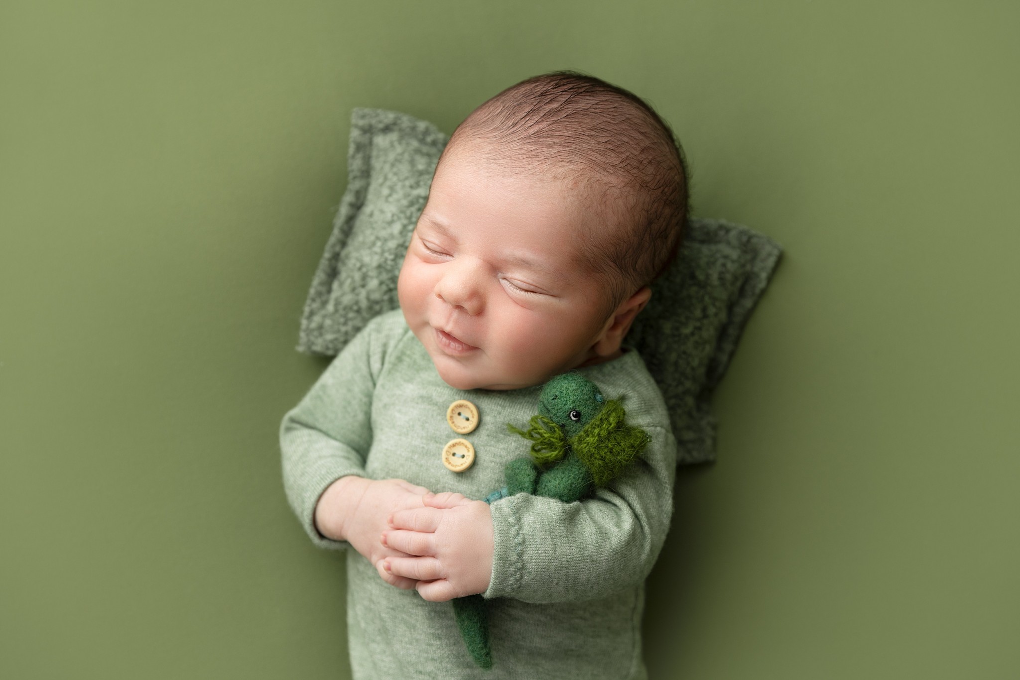 A happy newborn baby sleeps on a pillow while holding a knit green dinosaur