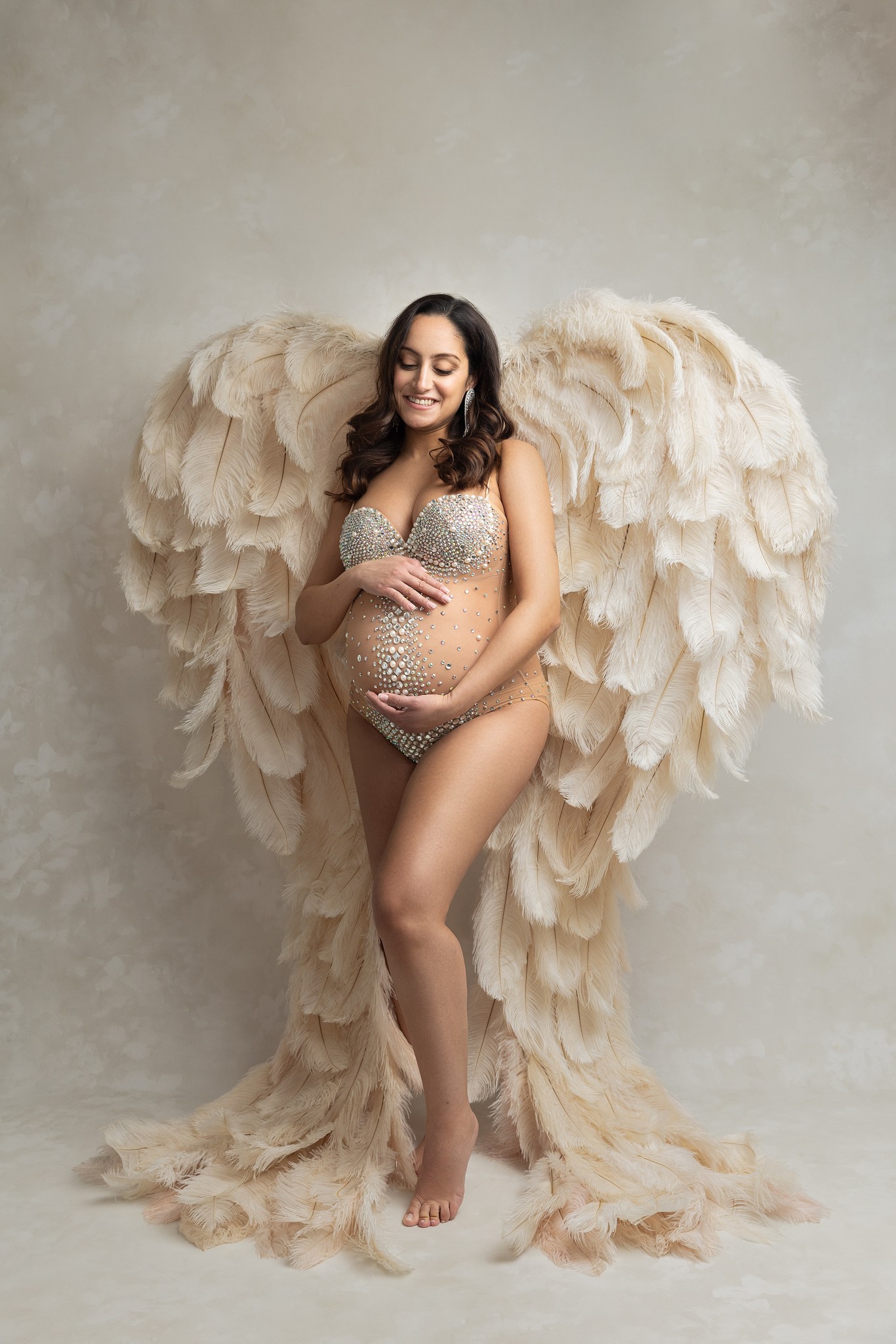 A mother to be in a jewel encrusted one piece stands in a studio with large feather angel wings
