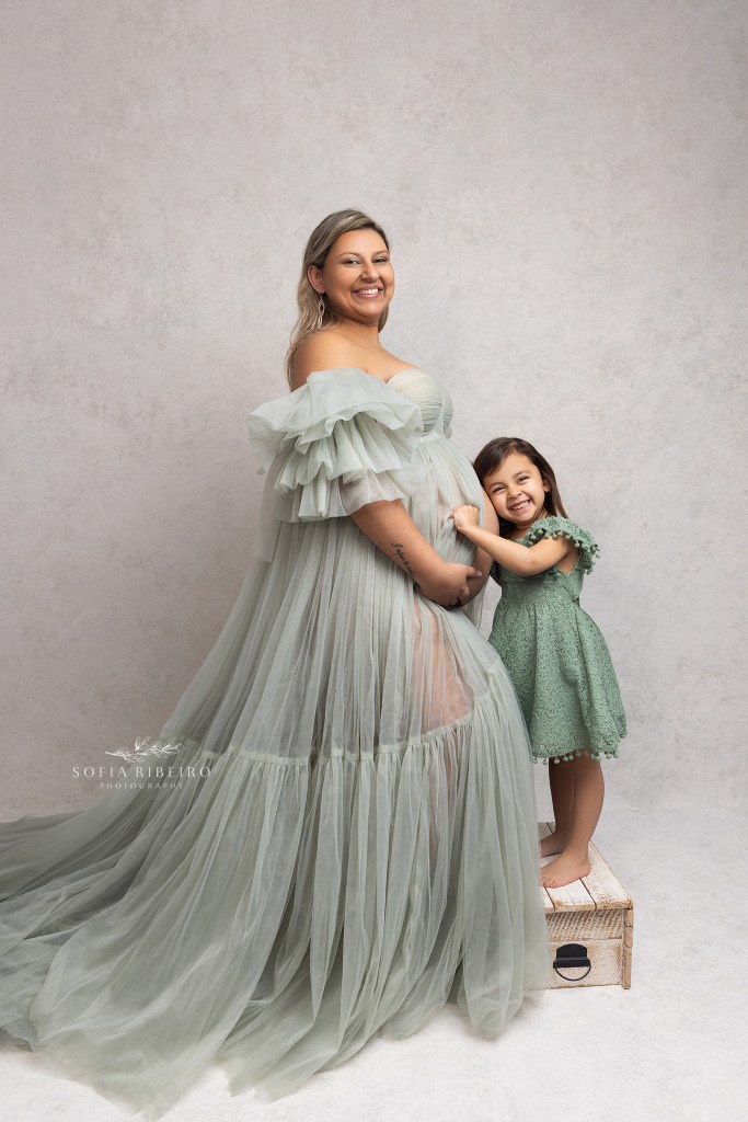 mom poses with her daughter holding her belly in matching green dresses to celebrate new baby in their maternity portraits