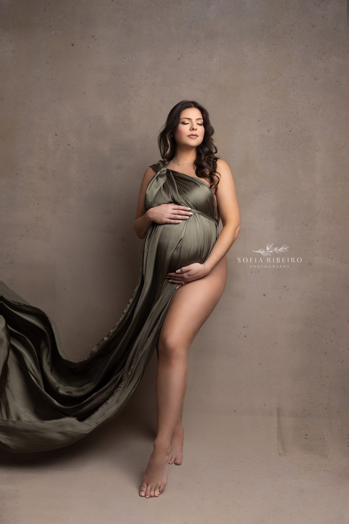 mom holds her baby bump while draped in an olive green flowing silk fabric during a maternity session in dover nj