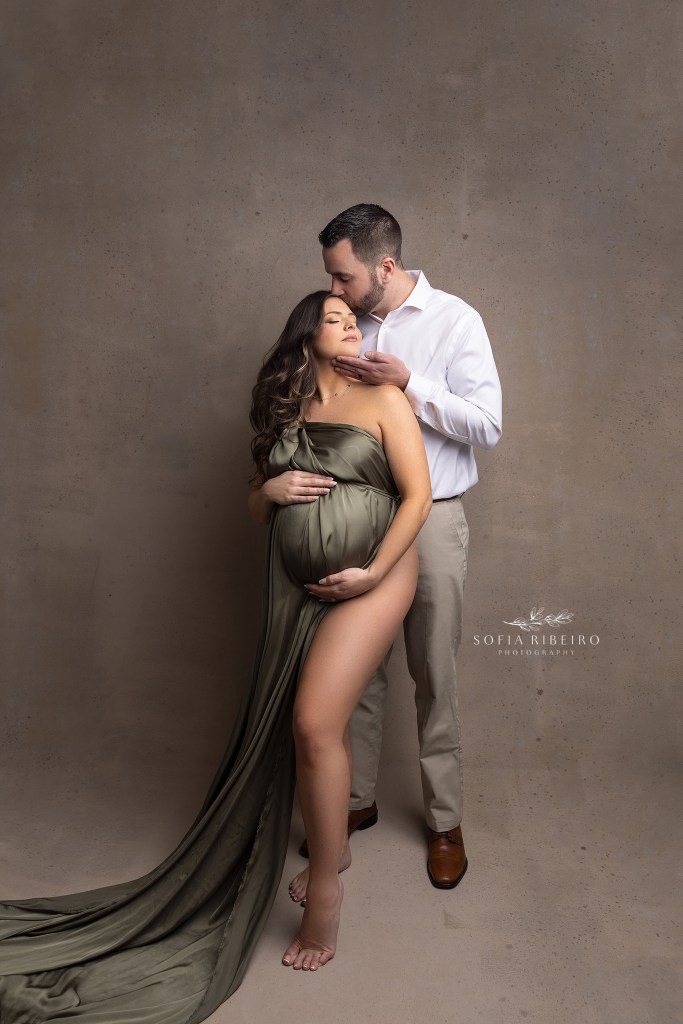 mom holds her baby bump while draped in an olive green flowing silk fabric as dad looks lovingly into her face