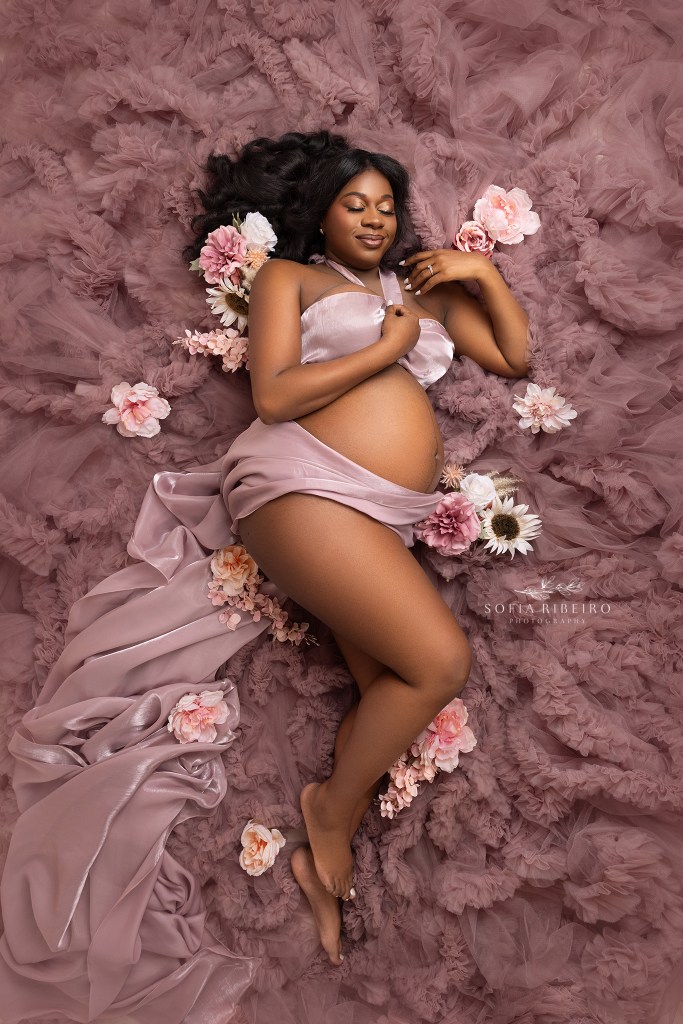 a mom poses in a bed of pink tulle, while draped in pink silk fabric meant to highlight her baby bump