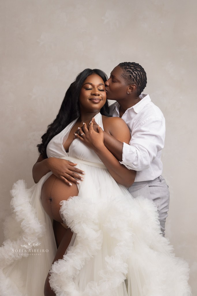 same sex couple poses together, sharing a sweet kiss on the cheek during their maternity session, dressed in white and neutrals during a session with an lgbtq maternity photographer in nj