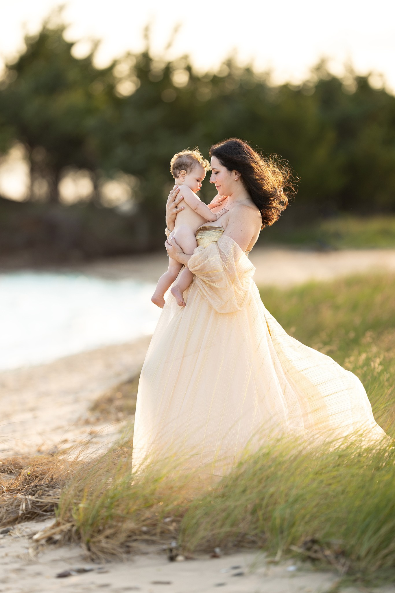 A mother cradles her infant baby against her chest while standing on a windy beach at sunset in a yellow dress