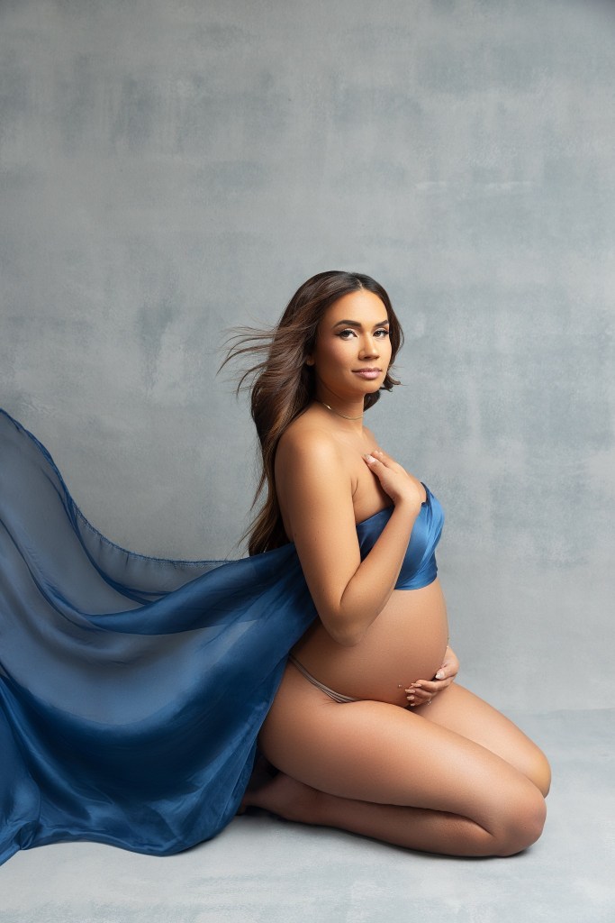 A mother to be kneels in a studio while covering with a flowing blue sheet