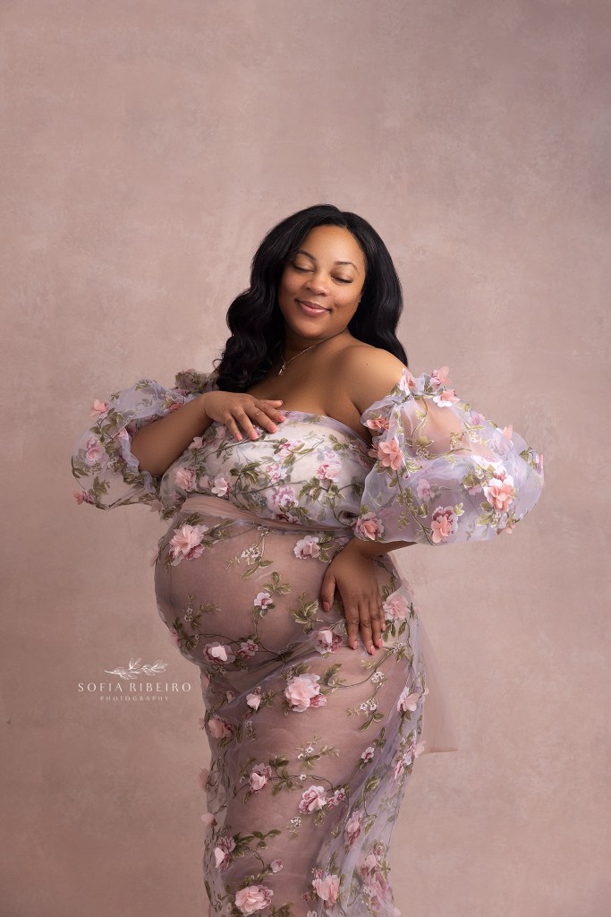 mom to be poses in a pink floral sheer dress during a maternity photo session in orange nj