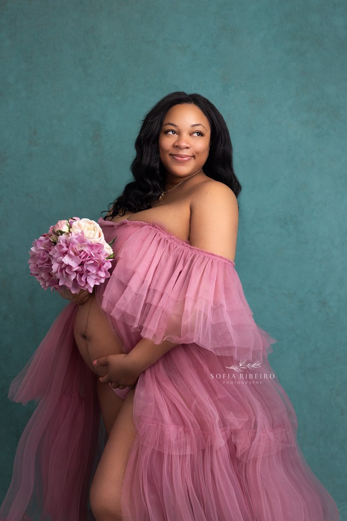 mom smiles while holding pink flowers and a gown celebrating her pregnancy with an orange nj maternity photographer