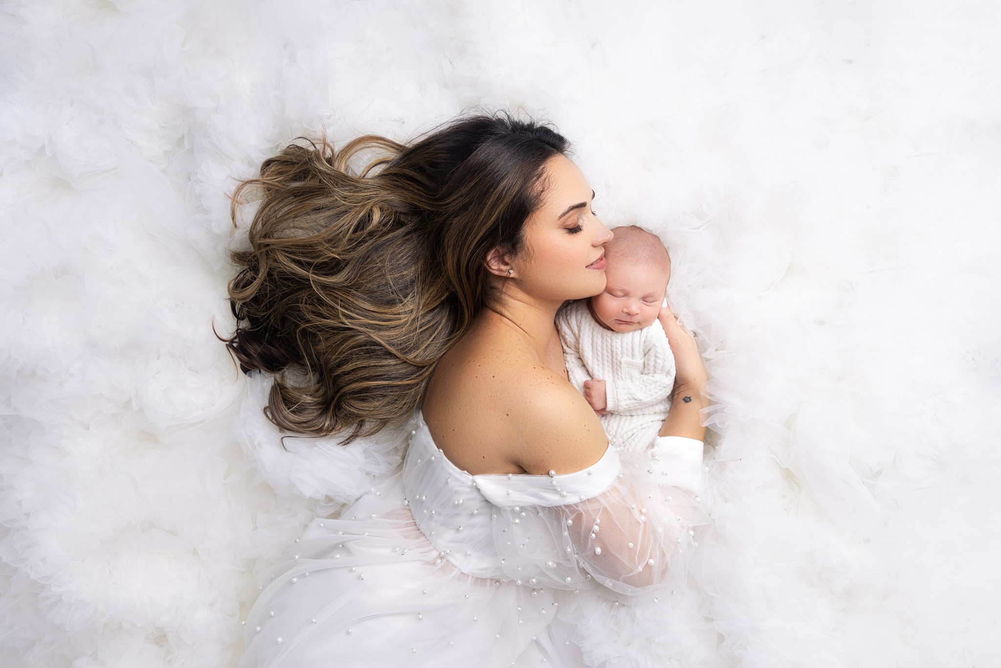A happy new mother in a white dress snuggles with her sleeping newborn baby in a white knit onesie after visiting a pediatrican middletown nj