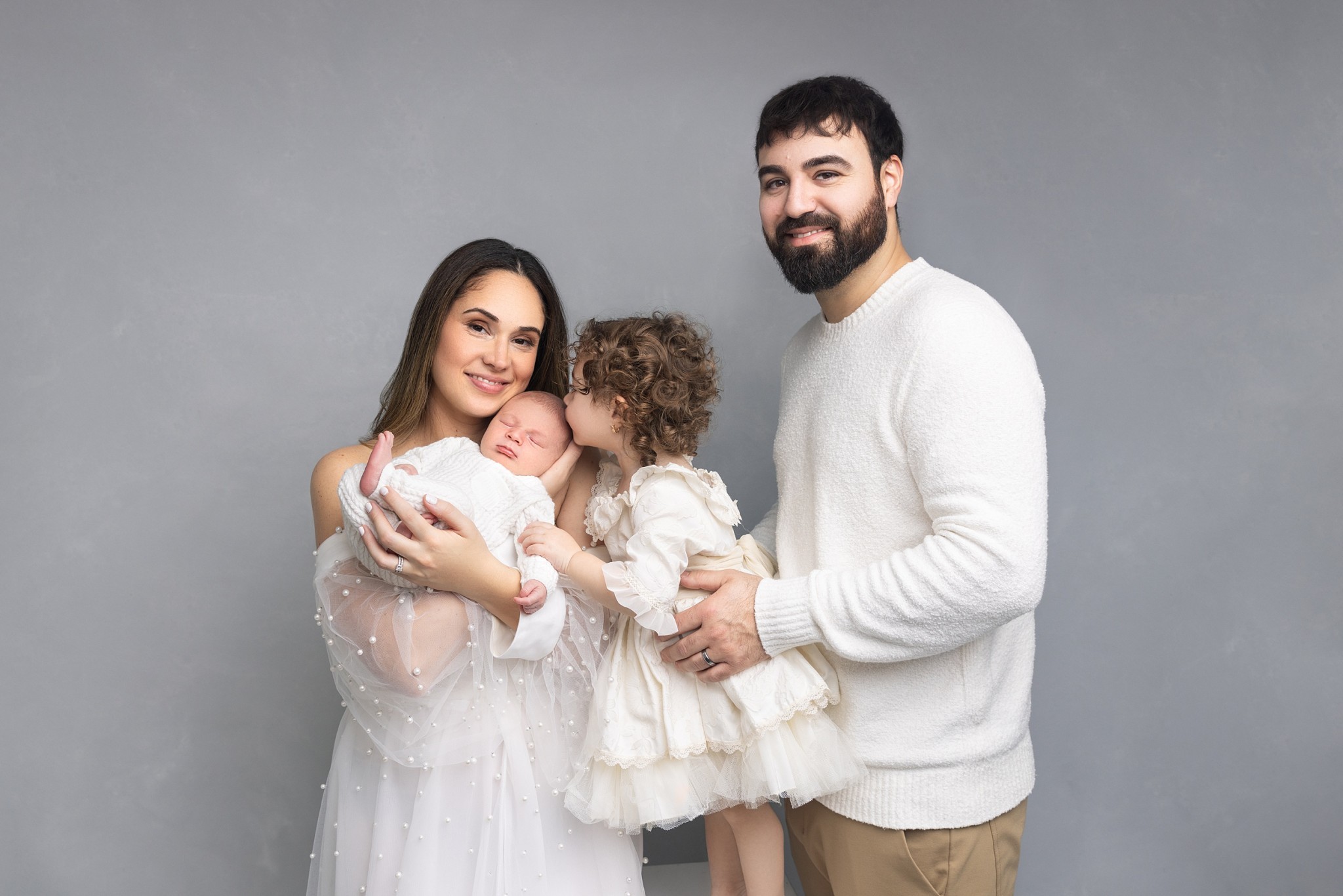 A mom and dad stand in a studio cradling their sleeping newborn as their toddler daughter kisses its forehead
