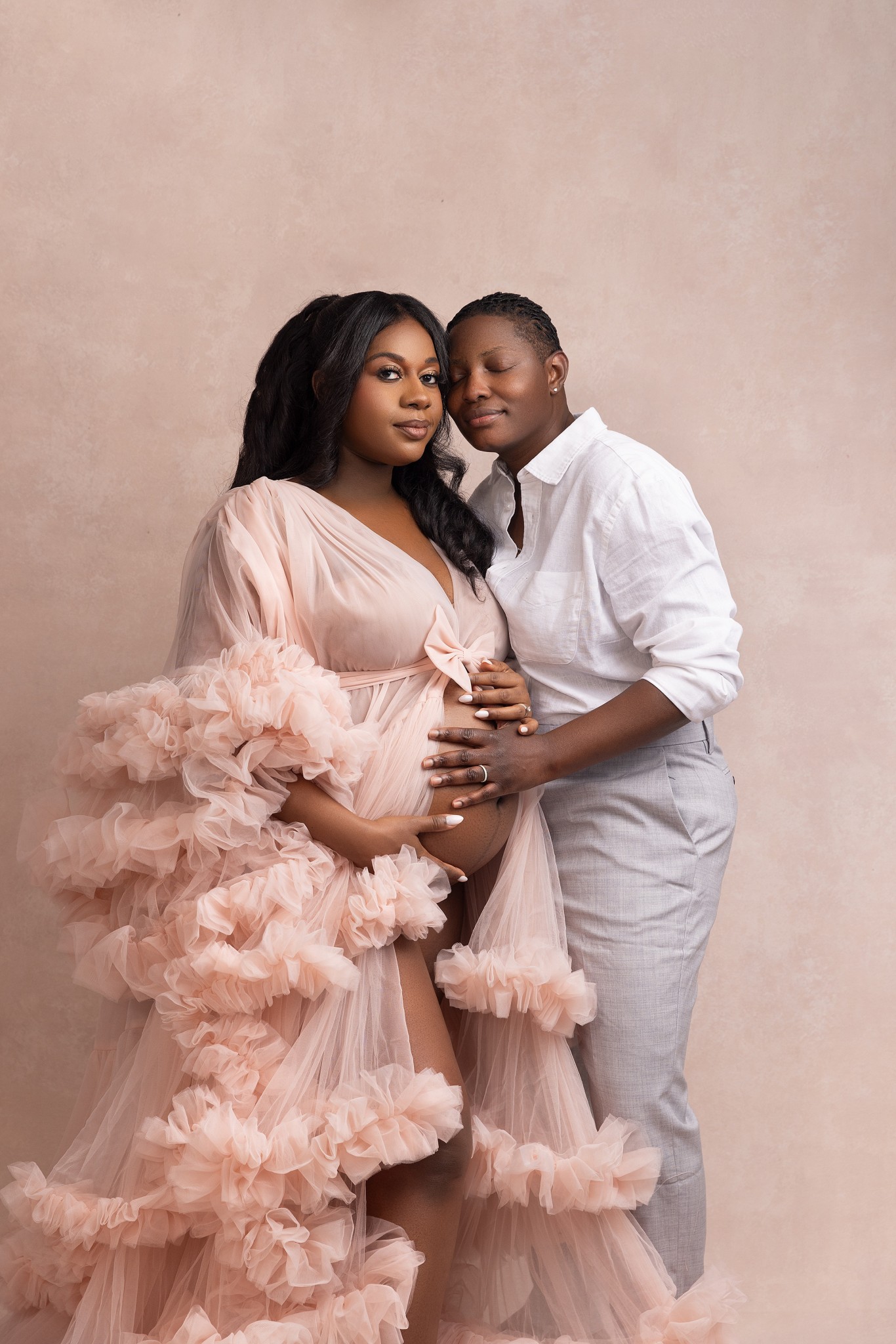 A woman in grey pants and white shirt snuggles her pregnant wife while standing in a studio holding the bump