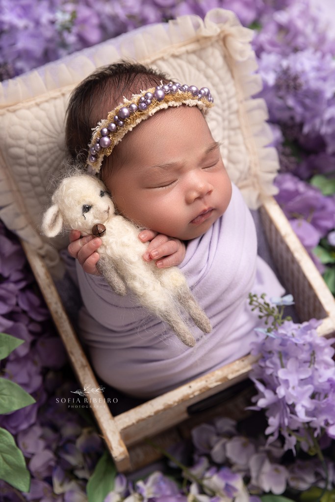baby girl holds a stuffed sheep snuggled in lilac flowers