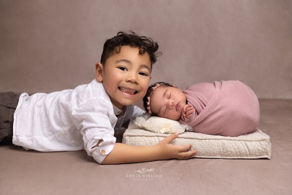 big brother snuggles his baby sister wrapped in a pink baby wrap and smiles