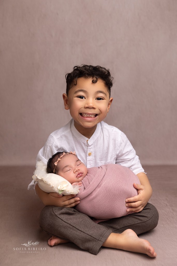 big brother snuggles his baby sister wrapped in a pink baby wrap and smiles