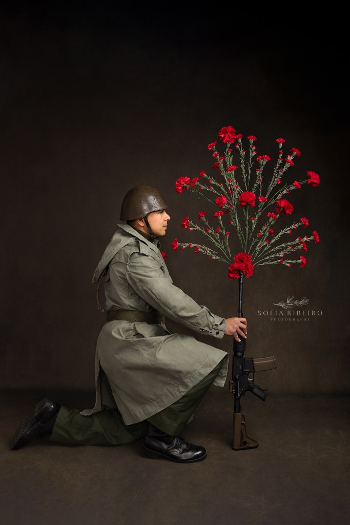 a model poses as a 70s styled solider, holding a rifle spraying carnations in every direction commemorating portugal's carnation revolution. Um modelo posa como soldado dos anos 70, com arma na mao a explodir com cravos comemorando o 50 aniversario da revolucao do dia 25 de abril em portugal
