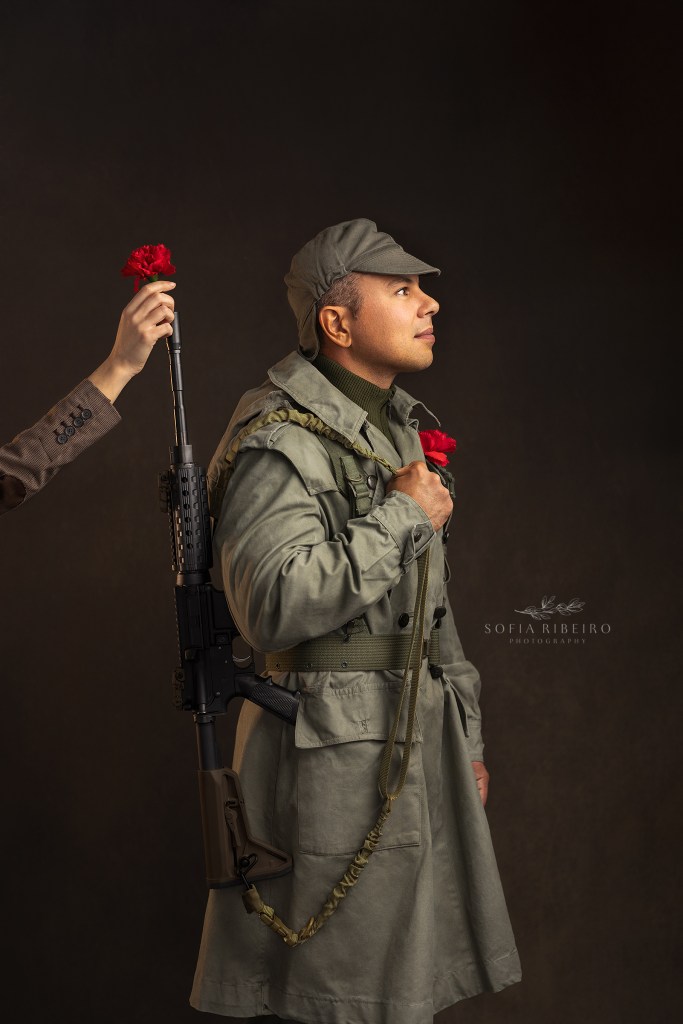 A hand places a carnation in the barrel of a gun on a 70s styled Portuguese soldier to commemorate the 25th of april's 50th anniversary of the carnation revolution in portugal. Um cravo e metido em uma arma em um retrato com estilo de tropa de anos 70, a celebrar o cinquentenario da revolucao de 25 de abril em portugal