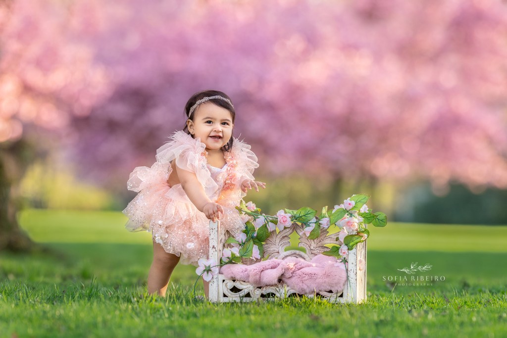 baby girl is smiling in a pretty pink dress with cherry trees in the background 