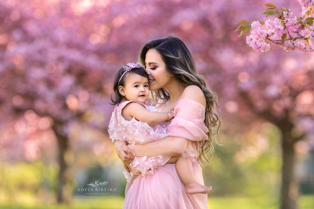 mom snuggles baby girl in the middle of cherry blossom blooms in nj