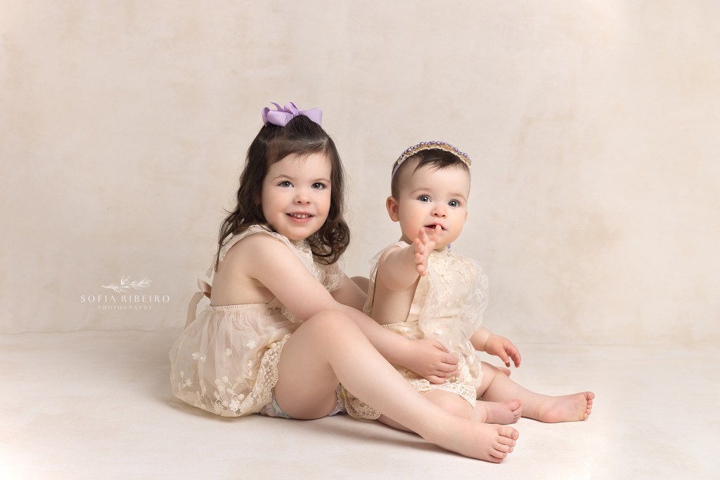 sisters pose in matching cream rompers during a baby session in westfield nj
