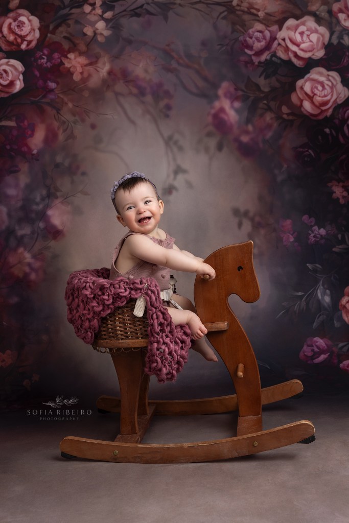 1 year old baby rocks on a vintage horse prop with a purple flower backdrop in westfield nj