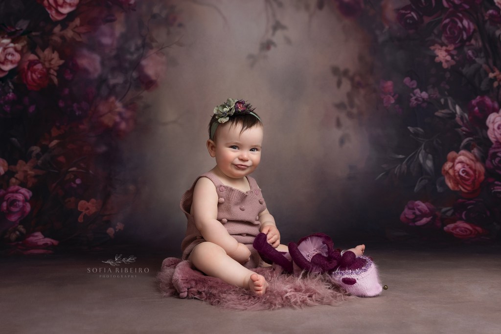baby girl sits against a floral backdrop during a aby portrait session, makes a silly face at the camera in westfield nj
