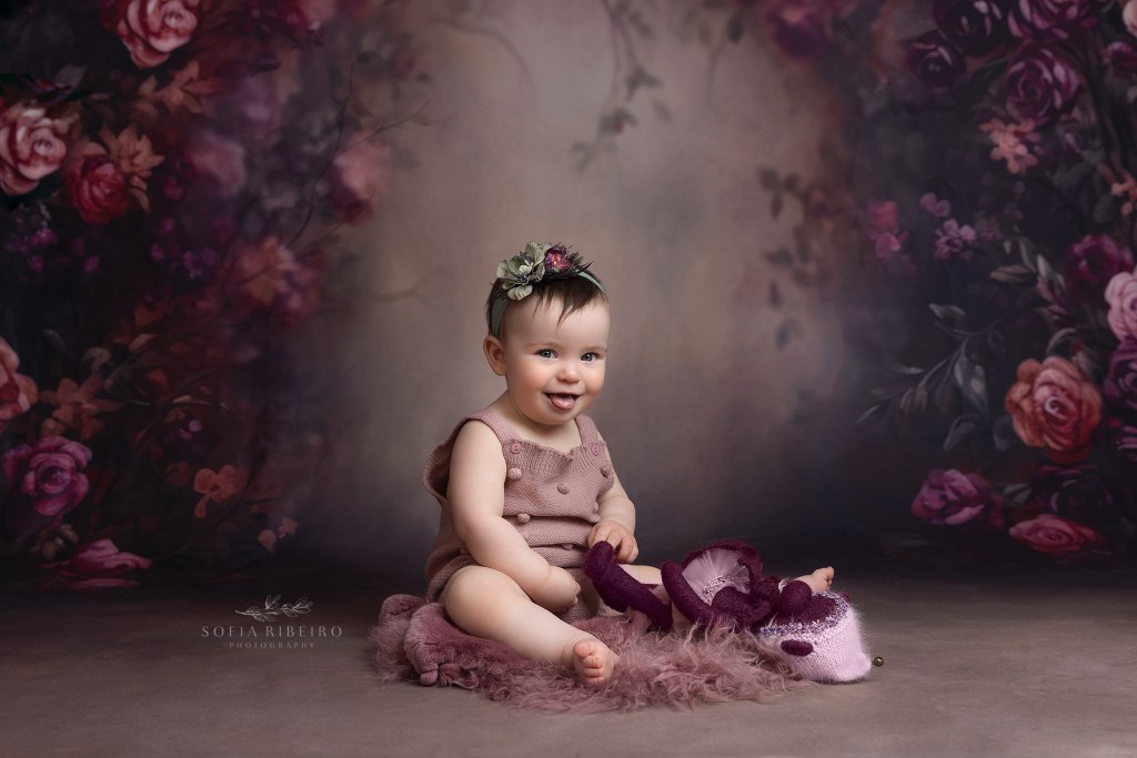 baby girl sits against a floral backdrop during a aby portrait session, makes a silly face at the camera in westfield nj