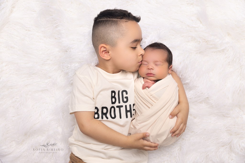 big brother kisses his new baby and wears a big brother tee while holding his new baby brother