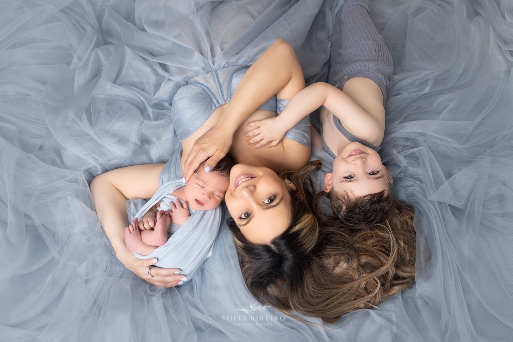 mom smiles surrounded by her babies during a photo session for her newborn