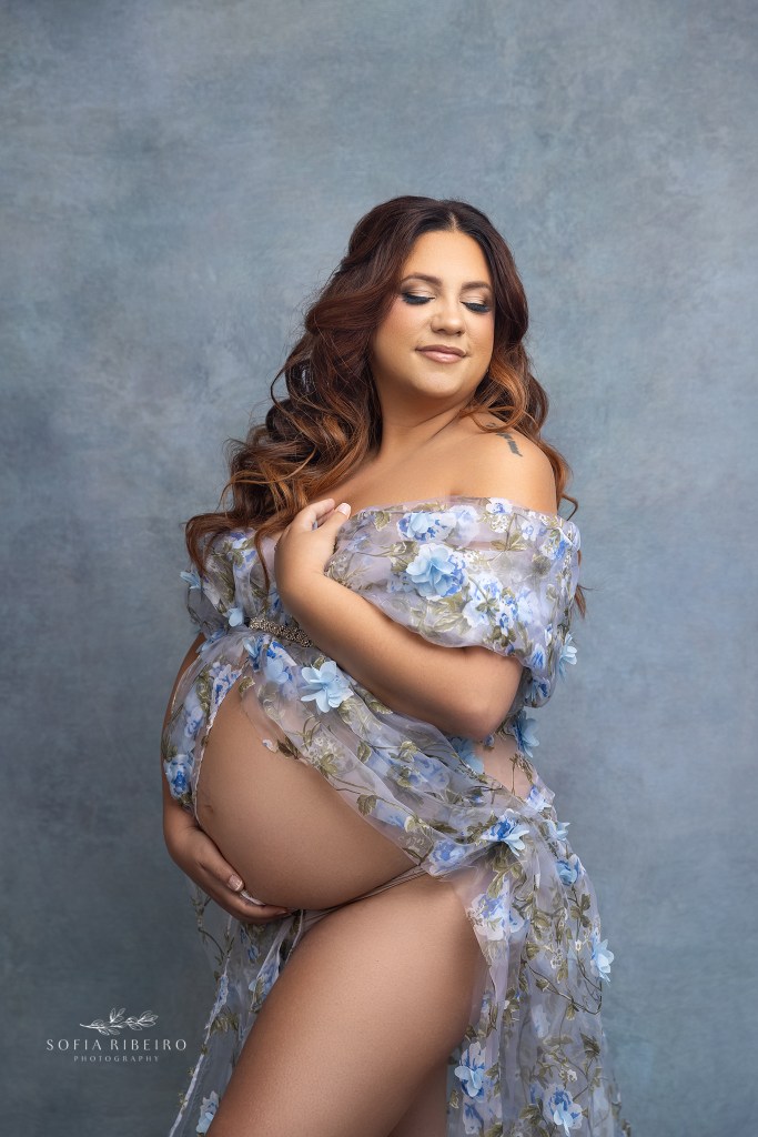 mom is draped in a sheer floral fabric in blue tones, while holding her baby bump snug