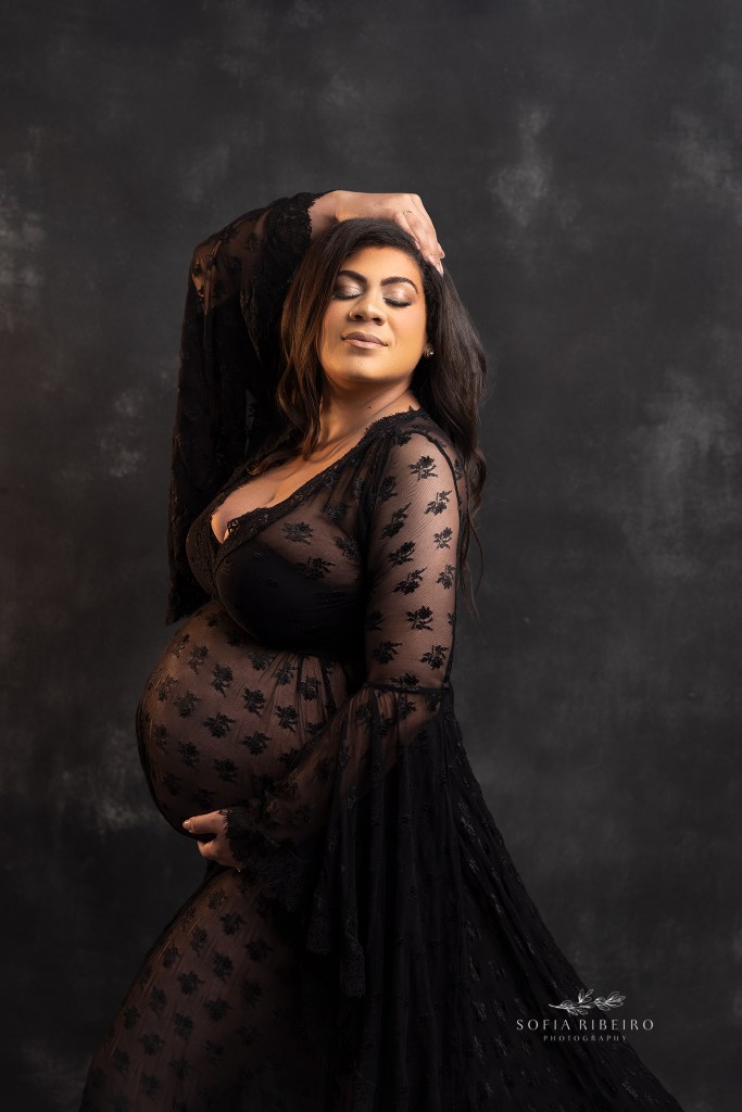 mom poses with her hand against her head, and another under her belly against a black backdrop for pregnancy photos