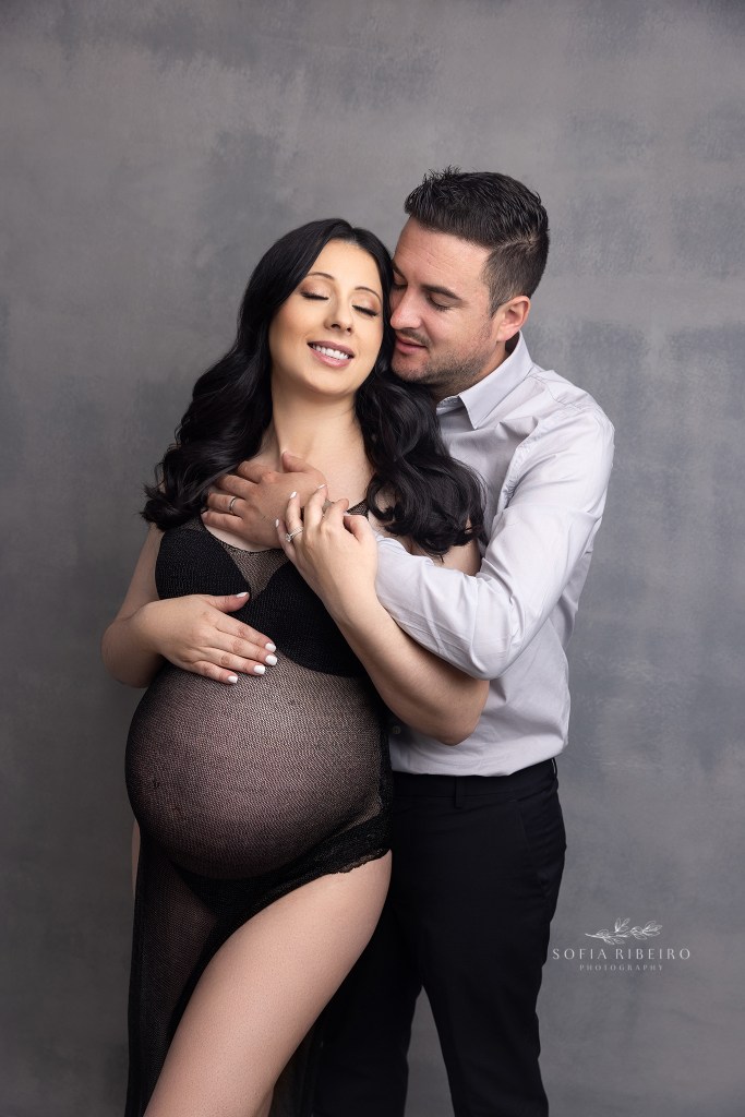 dad snuggles mom while placing a hand over her heart, showing the baby bump in a sheer black dress