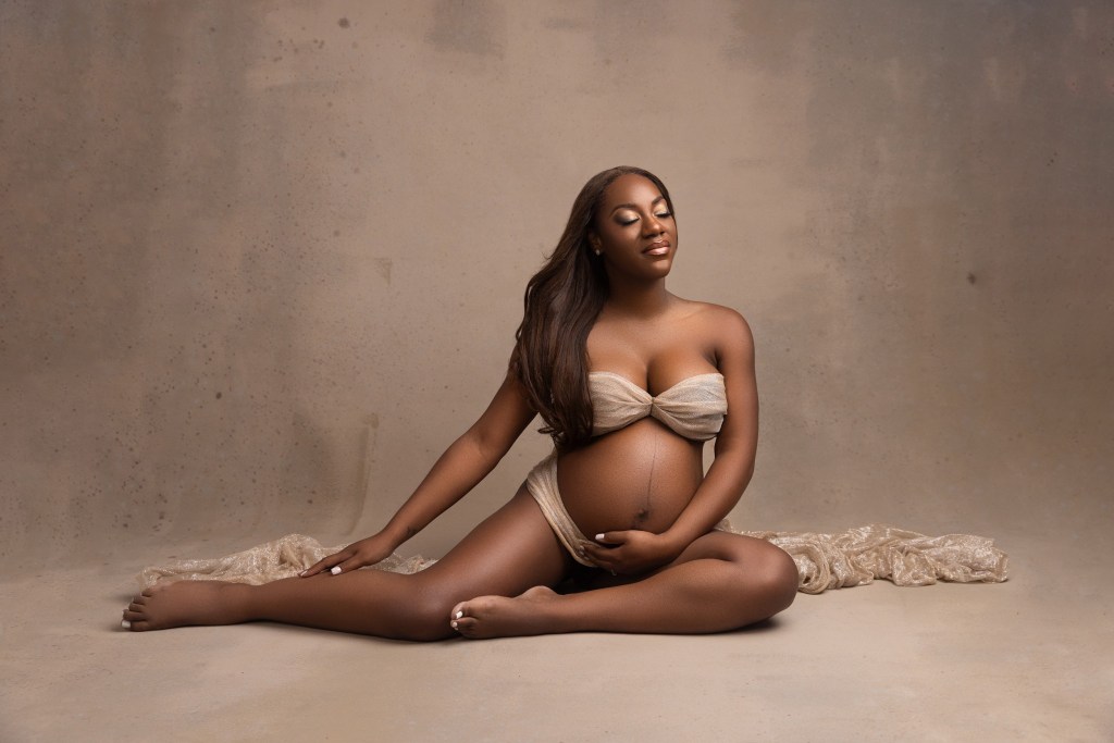 A mom to be wrapped in a fabric sits on the floor of a studio with a hand under her exposed bump before exploring baby shower venues union county nj