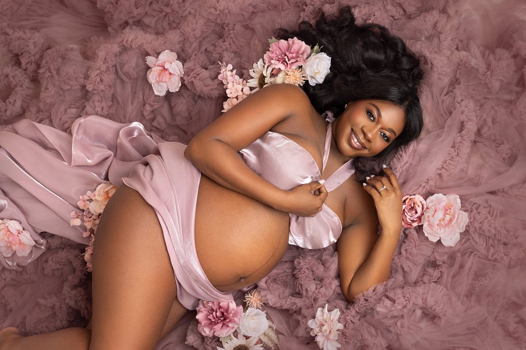 A pregnant woman lays on a bed of pink tulle during a photogra