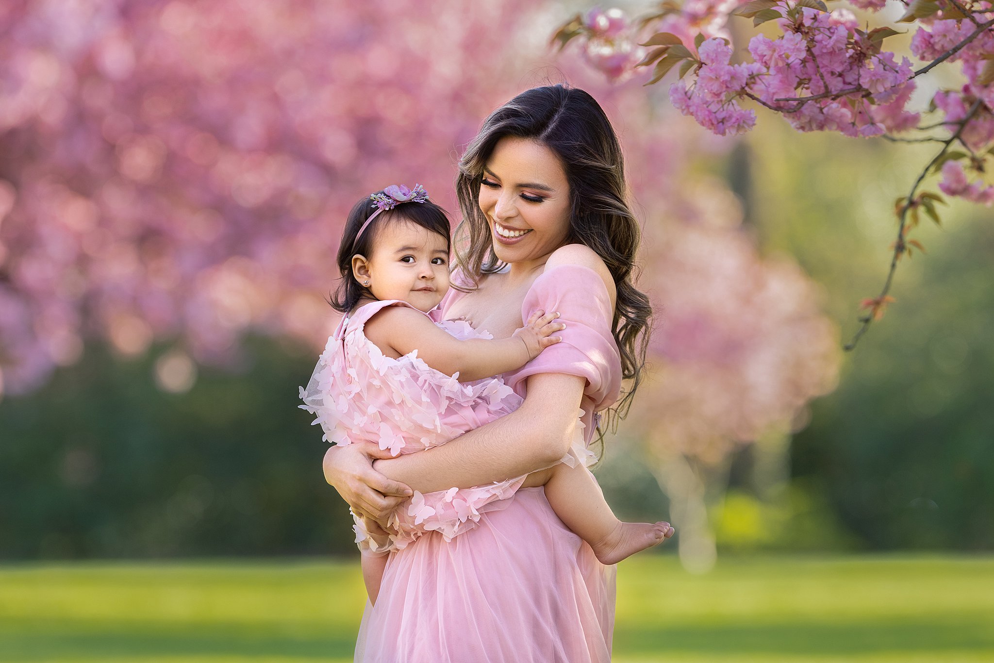 A smiling mother stands under a pink blooming tree in a matching dress with her toddler daughter on her hip