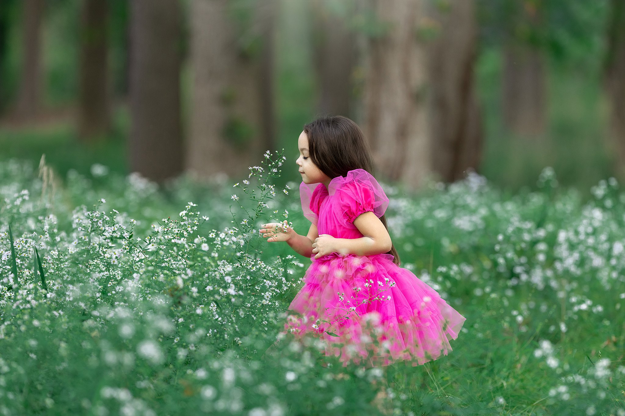 A young girl in a pink dress smells wildflowers in a forest