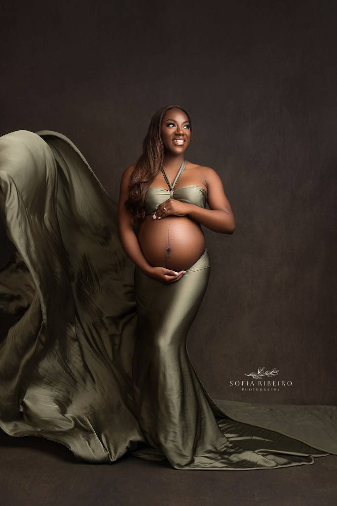 mom poses cradling her bump in green silk fabric, glowing during maternity photos in nj