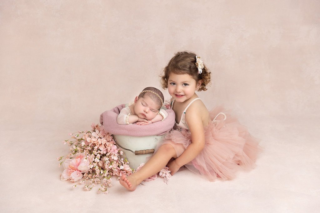 A toddler girl sits in a pink tutu in a studio with her sleeping baby sister in a metal bucket after visiting montclair baby